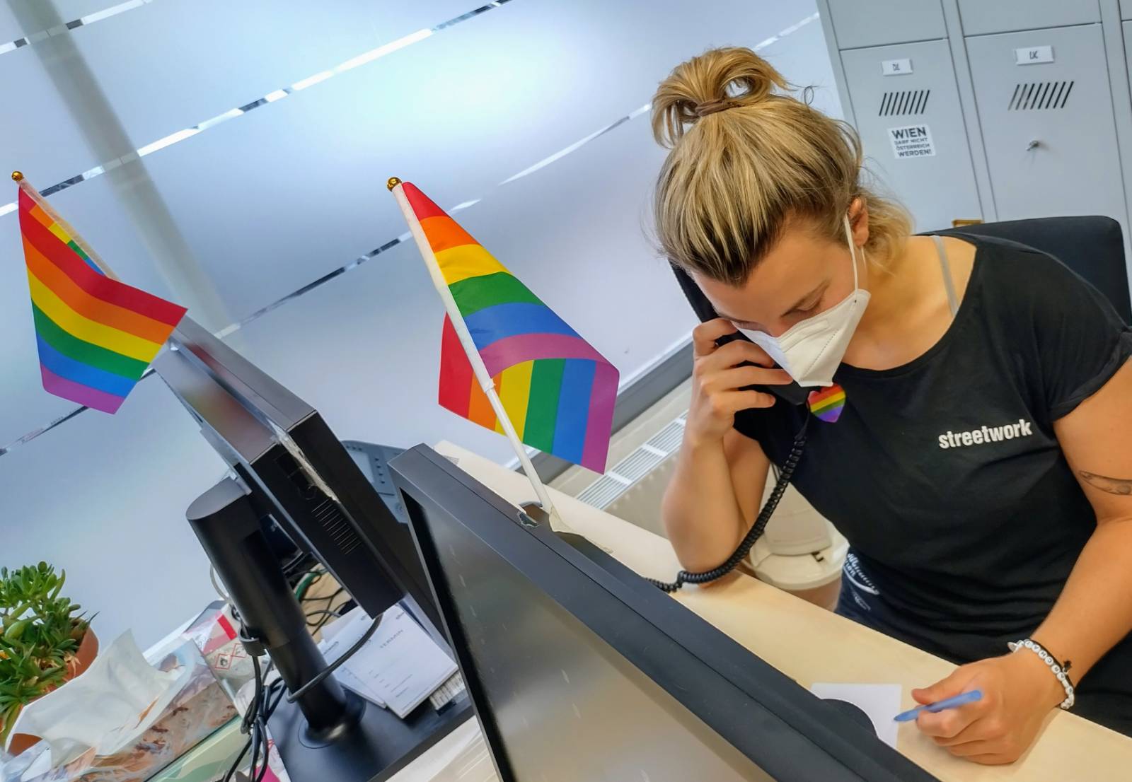 Mitarbeiterin von Streetwork sitzt vor ihrem PC Bildschirm und telefoniert. Am BIldschirm sind 2 Regenbogenfähnchen befestigt, sie trägt ein Regenbogen Herz Sticker auf der Kleidung.