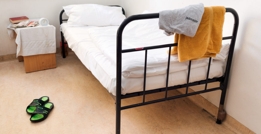 Bett in der Notschlafstelle – davor ein Paar Schlapfen, am Nachttisch persönliche Gegenstände