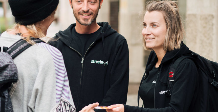 streetwork 2er-Team auf der Straße: ein Info-Folder wird einer Klient*in übergeben