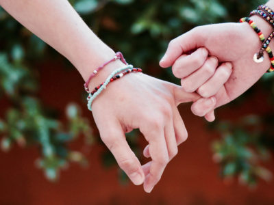 2 Hände mit vielen bunten Perlenbändern am Handgelenk – Händchenhalten über die beiden kleinen Finger