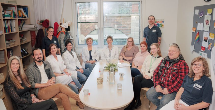 Gruppenbild im Ambulatorium: 13 Mitarbeiter*innen sitzen rund um einen Tisch